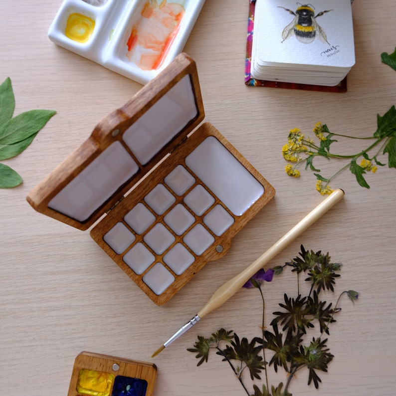 Puede incluir: Una paleta de pintura de acuarela de madera con 14 pozos vac&iacute;os, abierta y lista para usar. La paleta est&aacute; hecha de madera marr&oacute;n claro y tiene un interior blanco. Un pincel est&aacute; acostado junto a la paleta.