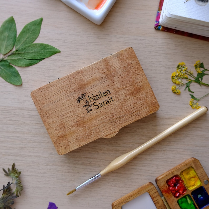 Puede incluir: Una caja de madera con tapa abatible, grabada con el texto "Nailea Sarait" y un dise&ntilde;o floral. La caja est&aacute; cerrada y se encuentra sobre una superficie de madera clara.