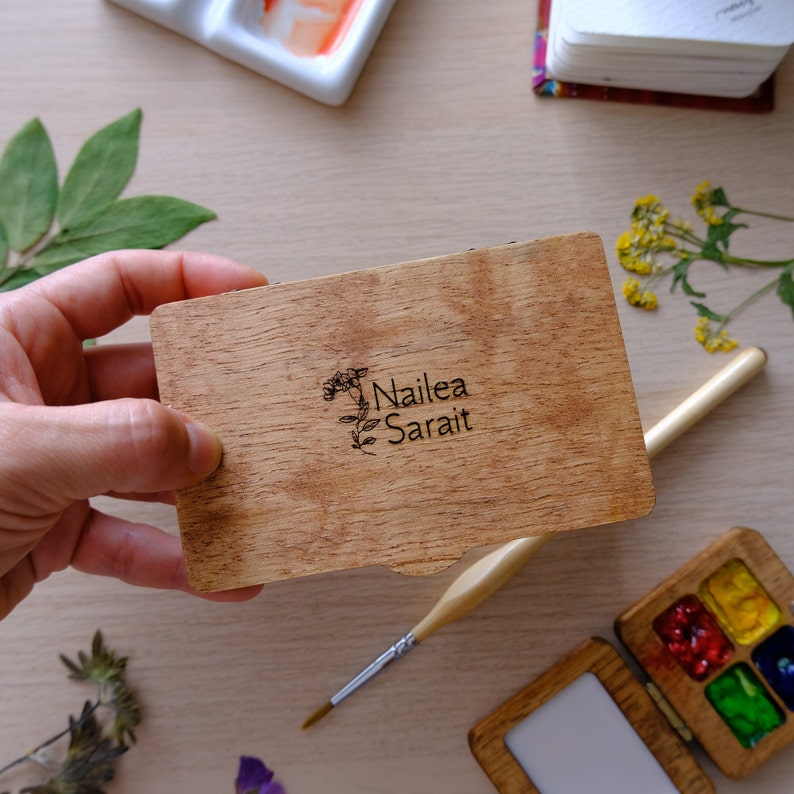 Puede incluir: Una caja de madera con tapa abatible, que muestra el texto "Nailea Sarai" y un dise&ntilde;o floral. La caja est&aacute; abierta y parece estar vac&iacute;a.