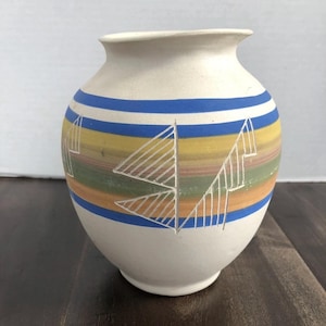 May include: A white ceramic vase with a rounded body and a narrow neck. The vase features horizontal stripes in blue, yellow, green, and orange. White geometric designs are etched into the painted surface. A decorative piece for the home.