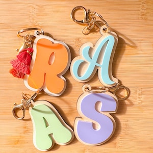 May include: Four colourful wooden letter keychains with gold metal rings. The letters are R, A, A, and S. The letters are in a retro style font. The keychain with the letter R has a red tassel.