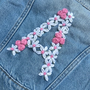 Puede incluir: Una chaqueta vaquera con un bordado floral blanco y rosa de la letra "A" en la espalda.