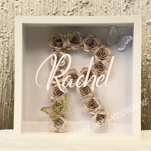 Könnte beinhalten: Ein weißer Rahmen mit einer Glasfront zeigt einen beigefarbenen Papierblumenbuchstaben "R" mit dem Namen "Rachel" in weißer Schrift. Zwei Schmetterlinge, einer silber und einer gold, sind ebenfalls im Rahmen enthalten.
