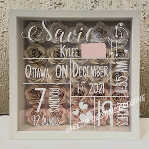 May include: A white shadow box with a glass front filled with paper flowers in shades of pink and beige. The box is decorated with white lettering that reads "Savile Kyler Ottawa, ON December 1st, 2021 1:35 AM 7 pounds 12 ounces @reina.designs.creative".