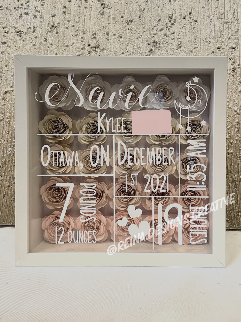 May include: A white shadow box with a glass front filled with paper flowers in shades of pink and beige. The box is decorated with white lettering that reads "Savile Kyler Ottawa, ON December 1st, 2021 1:35 AM 7 pounds 12 ounces @reina.designs.creative".