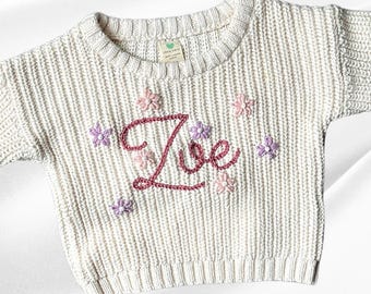 May include: A white knitted sweater with a pink and purple embroidered flower design and the name "Zoe" in pink thread.