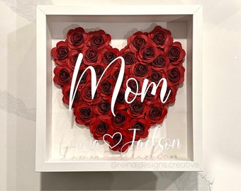 May include: White shadow box with a red paper flower heart that says "Mom" and "Jackson" with a small heart symbol. The bottom of the box says "@reina.designs.creative".
