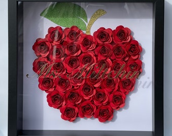 May include: A framed red paper flower apple with a green stem and gold glitter accents. The frame is black and the background is white. The text "Ms. Milch" is written in gold lettering.
