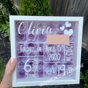 May include: A white shadow box with a purple flower design. The box includes the name Olivia, the date April 5th, 2020, the time 10:03 PM, and the baby's weight of 6 pounds and 5 ounces. The text "@Reina.Design.Creative" is also included.