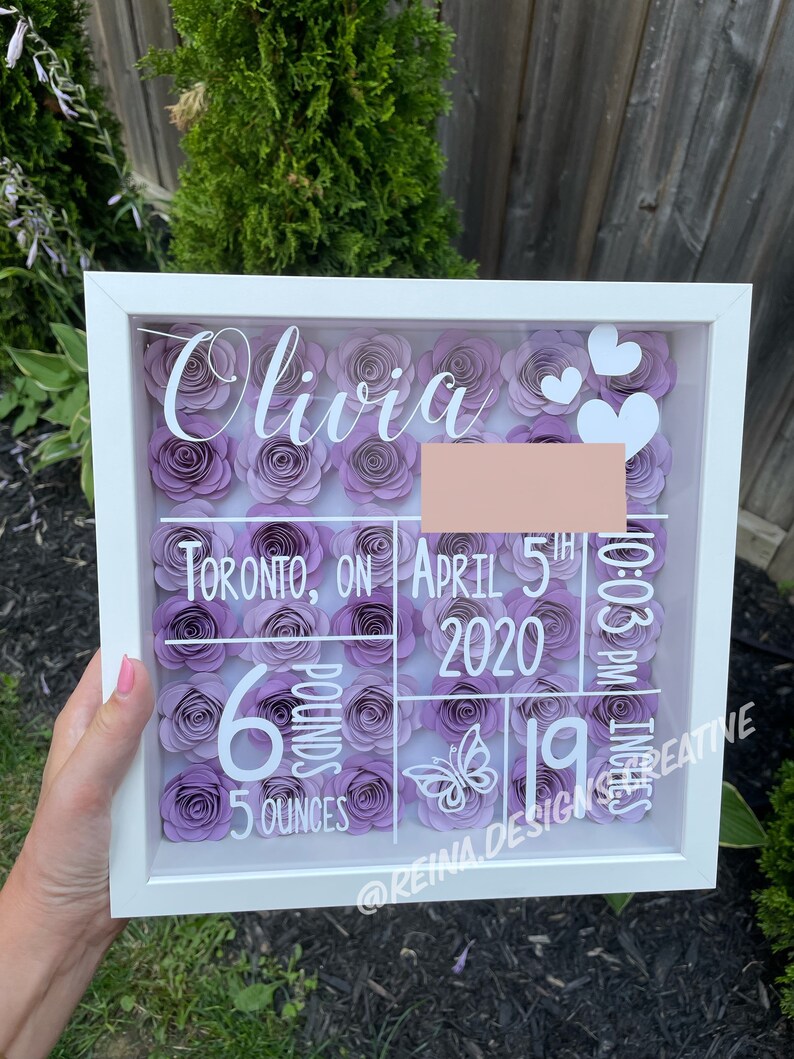 May include: A white shadow box with a purple flower design. The box includes the name Olivia, the date April 5th, 2020, the time 10:03 PM, and the baby's weight of 6 pounds and 5 ounces. The text "@Reina.Design.Creative" is also included.