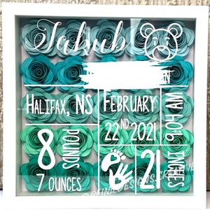 May include: A white shadow box with a teal and green paper flower background. The text "Jakub" is at the top of the box. The text "HALIFAX, NS FEBRUARY 22ND 2021" is in the middle of the box. The text "8 POUNDS 7 OUNCES" is in the bottom left corner of the box. The text "21 INCHES 9:04 AM" is in the bottom right corner of the box. A white footprint and handprint are in the bottom center of the box.