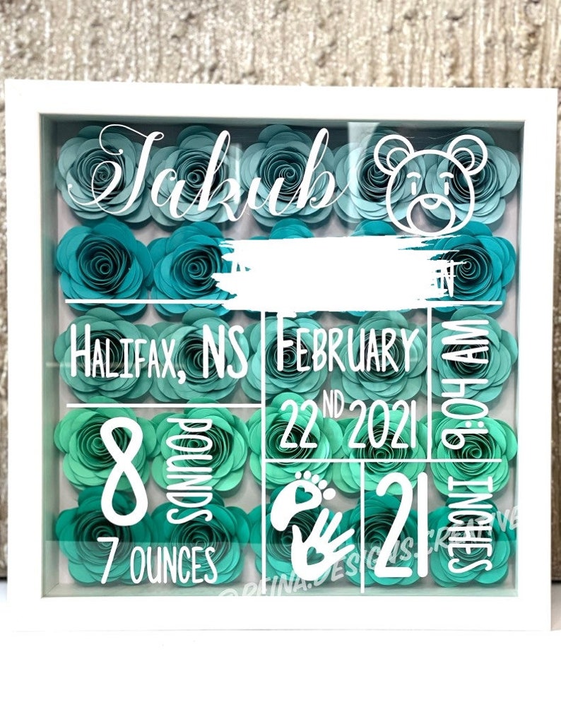 May include: A white shadow box with a teal and green paper flower background. The text "Jakub" is at the top of the box. The text "HALIFAX, NS FEBRUARY 22ND 2021" is in the middle of the box. The text "8 POUNDS 7 OUNCES" is in the bottom left corner of the box. The text "21 INCHES 9:04 AM" is in the bottom right corner of the box. A white footprint and handprint are in the bottom center of the box.