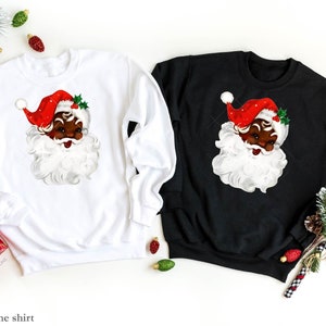 May include: Two Christmas sweatshirts, one white and one black, feature a vintage illustration of a Black Santa Claus with a red hat and white beard.