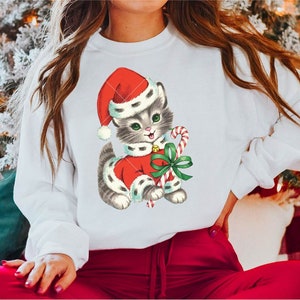 Puede incluir: Una sudadera blanca con una ilustraci&oacute;n vintage de un gatito gris que lleva un gorro de Pap&aacute; Noel rojo y sostiene un bast&oacute;n de caramelo. El gatito lleva un traje de Pap&aacute; Noel rojo y blanco con un lazo verde.