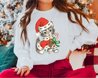 May include: A white sweatshirt featuring a vintage illustration of a grey kitten wearing a red Santa hat and holding a candy cane. The kitten is wearing a red and white Santa suit with a green bow.