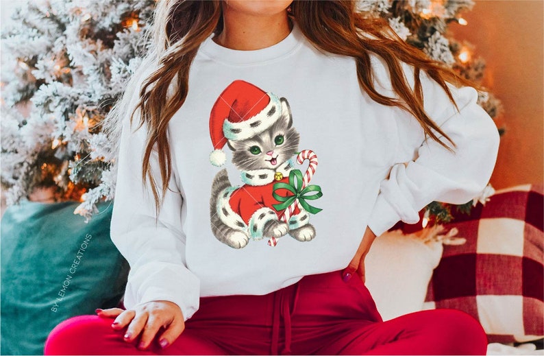 Puede incluir: Una sudadera blanca con una ilustraci&oacute;n vintage de un gatito gris que lleva un gorro de Pap&aacute; Noel rojo y sostiene un bast&oacute;n de caramelo. El gatito lleva un traje de Pap&aacute; Noel rojo y blanco con un lazo verde.