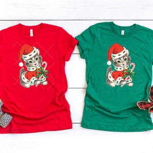 Puede incluir: Dos camisetas con un gatito navide&ntilde;o vintage que lleva un gorro de Pap&aacute; Noel y sostiene un bast&oacute;n de caramelo. La camiseta de la izquierda es roja y la camiseta de la derecha es verde.