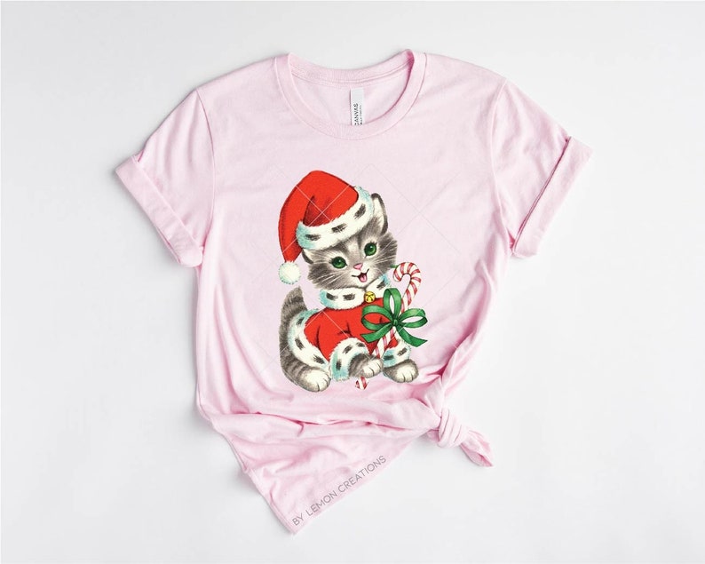 Puede incluir: Una camiseta rosa con una ilustraci&oacute;n vintage de un gatito gris y blanco con un gorro de Pap&aacute; Noel rojo y sosteniendo un bast&oacute;n de caramelo. El gatito lleva un traje de Pap&aacute; Noel rojo y blanco con un lazo verde.