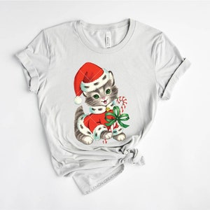 Puede incluir: Una camiseta gris con una ilustraci&oacute;n vintage de un gatito con un gorro de Pap&aacute; Noel y sosteniendo un bast&oacute;n de caramelo. El gatito lleva un traje de Pap&aacute; Noel rojo y blanco con un lazo verde.
