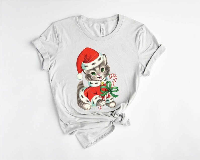 Puede incluir: Una camiseta gris con una ilustraci&oacute;n vintage de un gatito con un gorro de Pap&aacute; Noel y sosteniendo un bast&oacute;n de caramelo. El gatito lleva un traje de Pap&aacute; Noel rojo y blanco con un lazo verde.