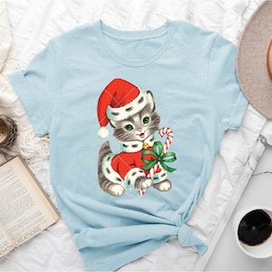 Puede incluir: Una camiseta azul claro con una ilustraci&oacute;n vintage de un gato con un gorro de Pap&aacute; Noel y sosteniendo un bast&oacute;n de caramelo. El gato lleva un traje de Pap&aacute; Noel rojo y blanco con un lazo verde.