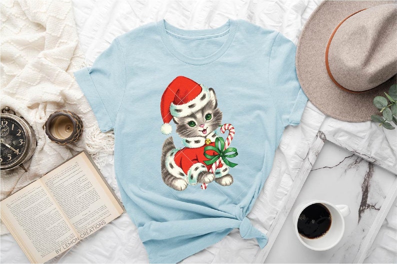 Puede incluir: Una camiseta azul claro con una ilustraci&oacute;n vintage de un gato con un gorro de Pap&aacute; Noel y sosteniendo un bast&oacute;n de caramelo. El gato lleva un traje de Pap&aacute; Noel rojo y blanco con un lazo verde.