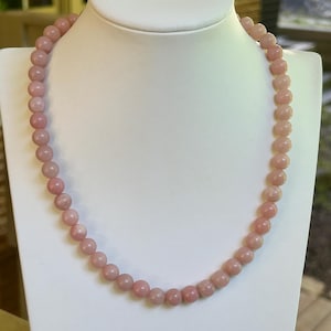May include: A pink beaded necklace displayed on a white jewelry bust. The necklace features round, light pink beads. The bust is set against a blurred outdoor background.