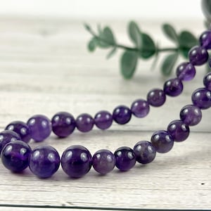 May include: A purple amethyst gemstone beaded necklace. The beads are round and smooth, and the necklace is strung on a clear thread.
