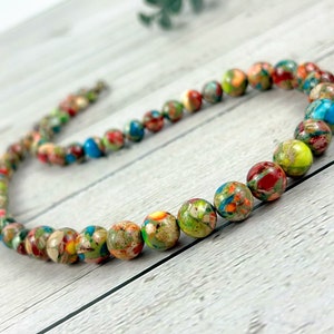 May include: A colorful beaded necklace made of multi-colored jasper stones. The necklace is made of round beads with a speckled pattern of green, red, yellow, and blue.