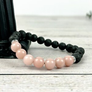 May include: A bracelet made with black lava rock beads and pink gemstone beads. The bracelet is on a light wood surface.