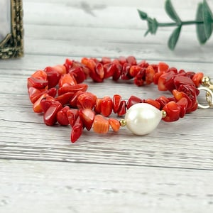 May include: A red coral bracelet with a large white pearl bead. The bracelet is made of small, irregularly shaped coral pieces and has a gold-colored clasp. The jewelry is displayed on a light-colored wooden surface.