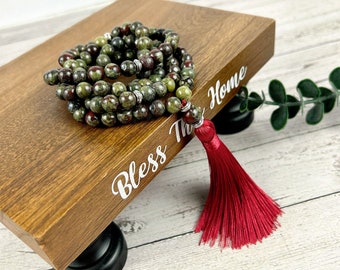 May include: A green and red gemstone mala with a red tassel. The mala is on a wooden stand with the words "Bless This Home" written on it.