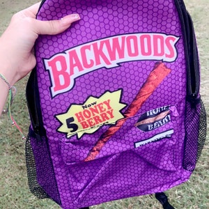 May include: Purple backpack with a honeycomb pattern and the Backwoods logo. The backpack features a front pocket with a graphic of a rolled tobacco product and the words "Now Honey Berry". Black mesh side pockets and black straps.