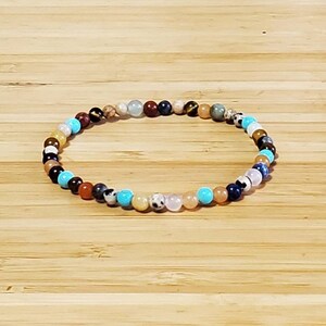 May include: A beaded bracelet featuring a variety of small, colorful gemstones. The bracelet includes beads in shades of blue, brown, white, black, and tan. The bracelet is displayed on a light brown wooden surface.