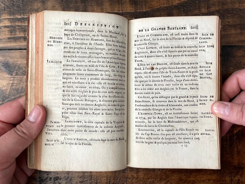May include: An open book with yellowed pages and a brown leather cover. The book is open to a page with the title "Description" and text in French about the island of Jamaica. The next page is titled "De la Grande Bretagne" and contains text about the island of Cumberland.