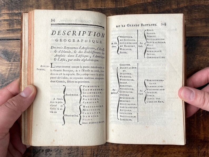 May include: An open book with a black and white text on a white background. The text is in French and describes the geography of England, Ireland, Scotland, Africa, America, and Asia. The book is open to pages 1vj and 1vij.