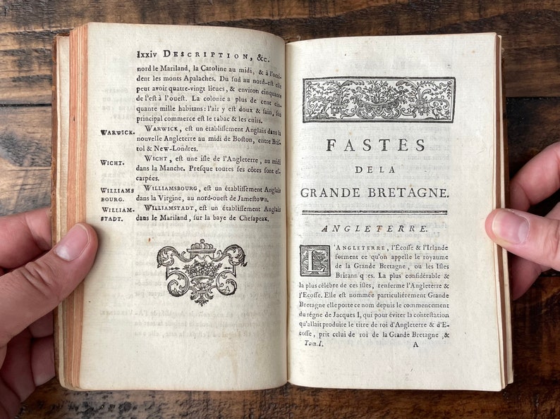 May include: An open book with a title page that reads "Fastes de la Grande Bretagne." The book is open to a page with text in French about the history of England, Scotland, and Ireland. The book is bound in brown leather and has a decorative design on the cover.