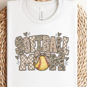 May include: A white t-shirt featuring the text SOFTBALL MOM in a mix of leopard print, camouflage, and checkered patterns. A yellow softball with red stitching and a leopard print bow is centred in the design, accented by small heart icons.