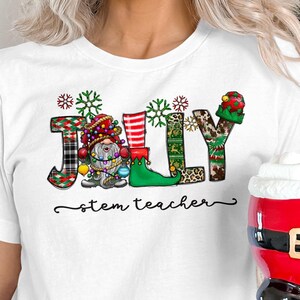 Puede incluir: Camiseta blanca con la palabra "JULLY" escrita en letras de colores con patrones. Cada letra está decorada con un diseño diferente de gnomo navideño. El texto "stem teacher" está escrito debajo de la palabra "JULLY".