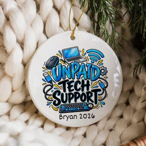 May include: A round white ceramic ornament featuring a colorful graphic of a laptop, mouse, keyboard, and cables. The text reads UNPAID TECH SUPPORT and Bryan 2026. The ornament hangs from a gold string against a white knitted background.