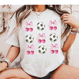 May include: White t-shirt with a repeated pattern of pink bows, cherries, and soccer balls.