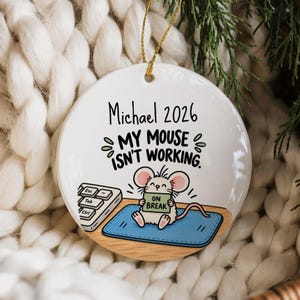 Puede incluir: Un adorno redondo de cerámica blanca con un ratón de dibujos animados sentado en una alfombrilla azul junto a un teclado. El ratón sostiene un cartel que dice ON BREAK. El texto superior dice Michael 2026 y MY MOUSE ISN'T WORKING. Cordón dorado.