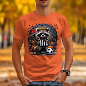 May include: Orange t-shirt featuring a cartoon raccoon referee holding a red card and standing next to a soccer ball. The raccoon is wearing a black and white striped shirt. The background includes pumpkins, bats, and a spooky tree design.
