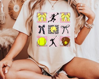 May include: Cream-colored t-shirt featuring a grid of softball-themed graphics. The design includes yellow bows, softball silhouettes, a softball, a glove, and the text "GAME DAY". The shirt has a relaxed fit.