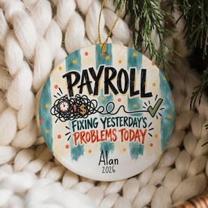 以下が含まれることがあります： ティールとクリーム色のストライプの背景に「PAYROLL」、「FIXING YESTERDAY'S PROBLEMS TODAY」、「Alan」、「2026」の文字が書かれた円形のセラミック製オーナメント。時計、ドル記号、緑色のチェックマークのグラフィックと金色の紐が付いています。
