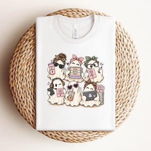 Peut inclure: Un t-shirt blanc avec un motif de six fantômes de dessin animé portant des bandeaux et des accessoires. Les fantômes tiennent des objets comme une tasse de café, une pile de livres étiquetés Payroll Benefits Reports, un ordinateur et une calculatrice.