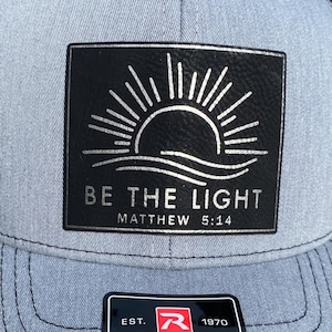 May include: A grey and black trucker hat featuring a rectangular black patch with a white graphic of a rising sun over waves. The text reads BE THE LIGHT MATTHEW 5:14. A sticker on the brim displays the text RICHARDSON EST. 1970 ORIGINAL 112 TRUCKER.