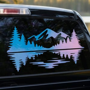May include: A decal applied to a truck rear window featuring a mountain range and pine forest silhouette with a gradient color effect transitioning from blue to pink, including a mirrored reflection of the landscape on the water surface below.