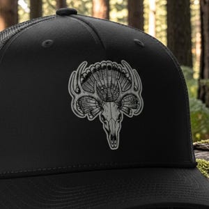 May include: A black trucker hat featuring a gray graphic of a deer skull with antlers integrated with a turkey fan. The hat is constructed with a solid black front panel and a charcoal mesh back. The text on the image reads: flint/black leatherette black/charcoal mesh hat.