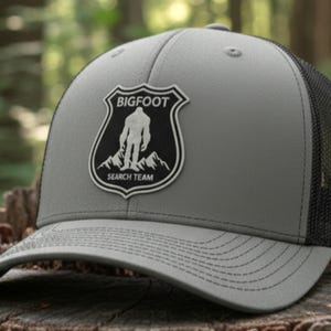 May include: Gray trucker hat with a black mesh back and a curved brim. The hat features a Bigfoot Search Team patch with a silhouette of Bigfoot and mountain graphics. The hat is made of charcoal and black materials.